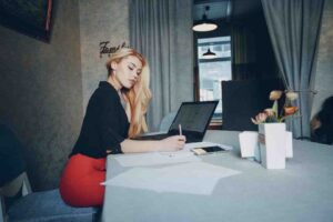 A woman in a black blouse and red pencil skirt writes while seated at a table with a laptop