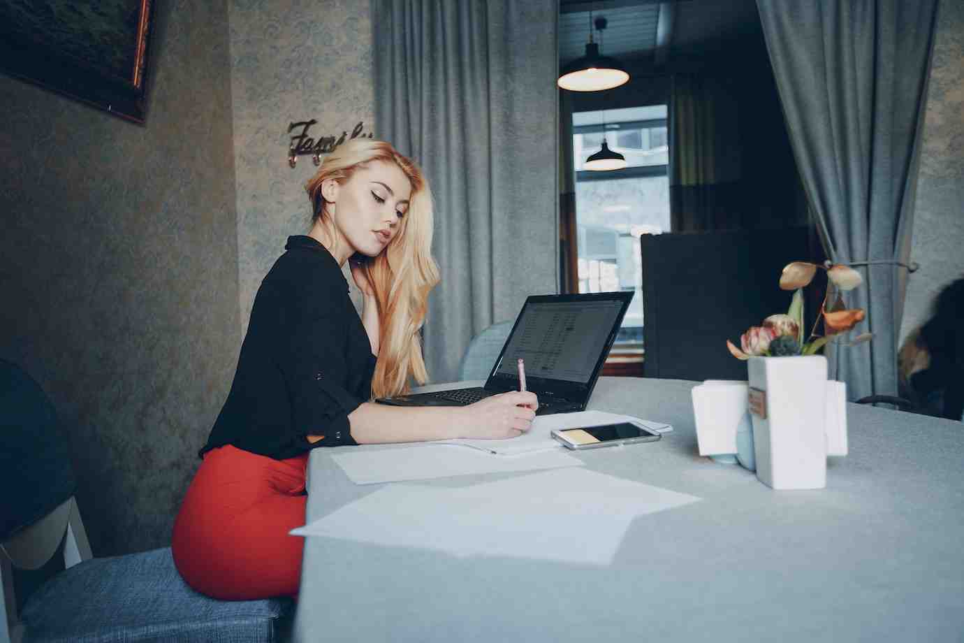 A woman in a black blouse and red pencil skirt writes while seated at a table with a laptop