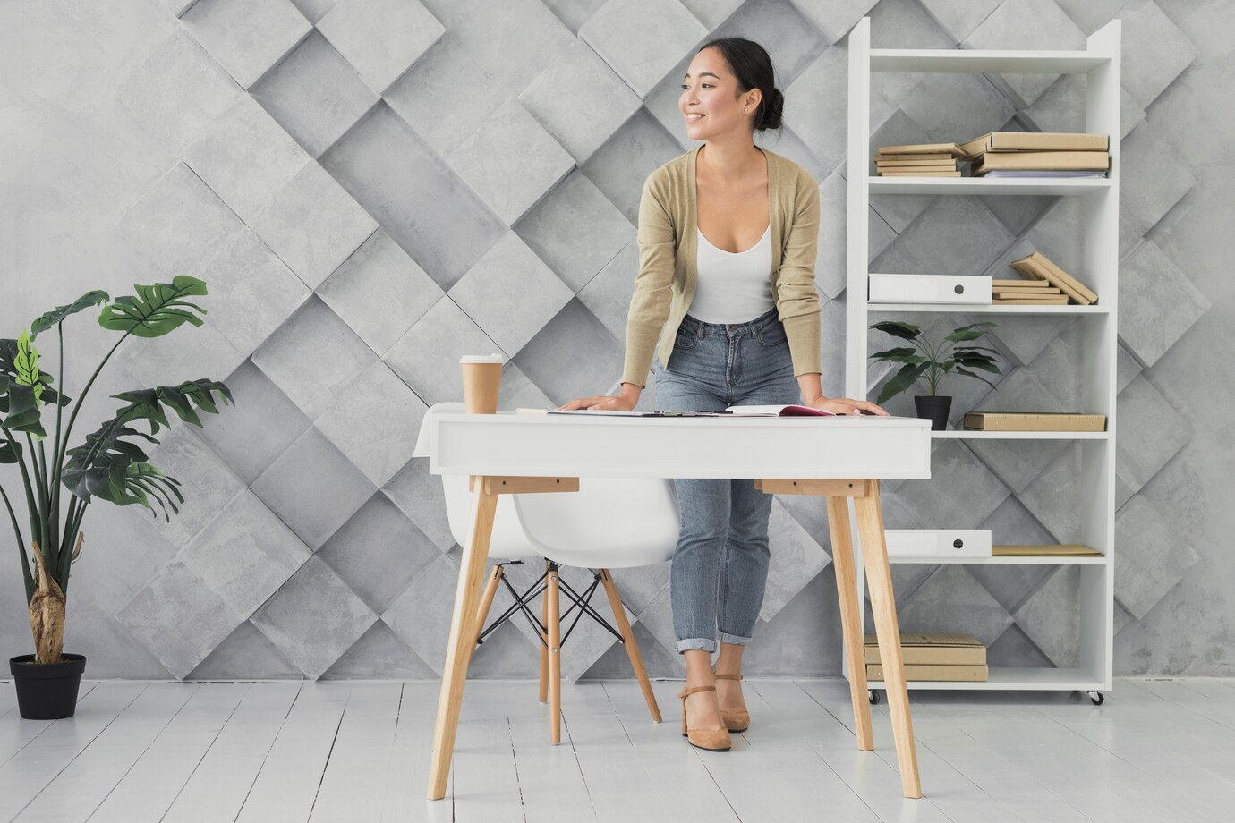 A modern workspace featuring a white desk with books, a coffee cup, and a potted plant, against a geometric gray wall and shelving unit.