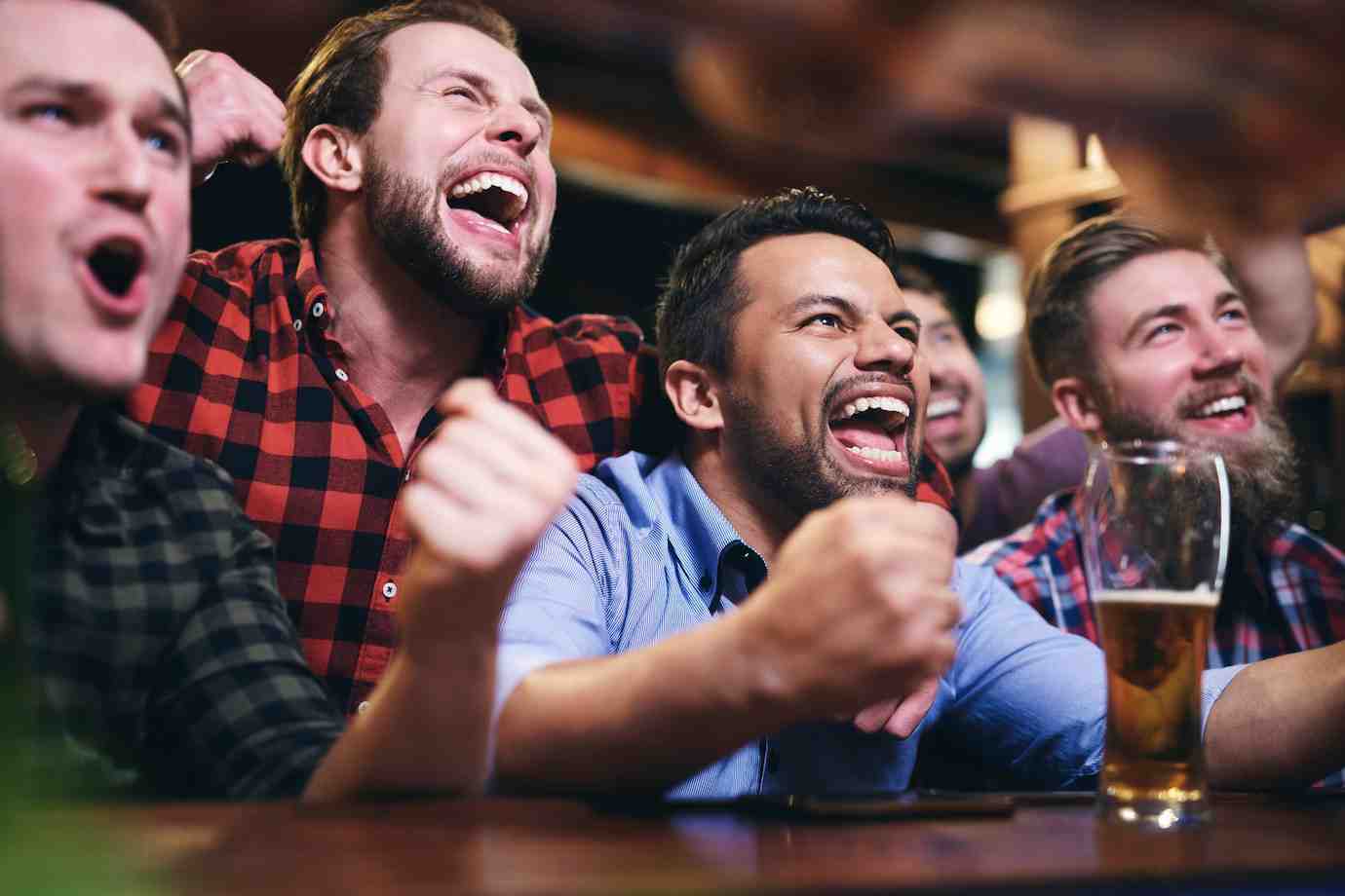 A group of friends celebrates at a bar, showing excitement with raised fists and enjoying drinks together.