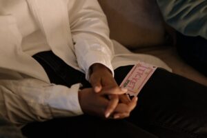 A person wearing a white shirt sits with clasped hands and a colorful ticket resting on their thigh
