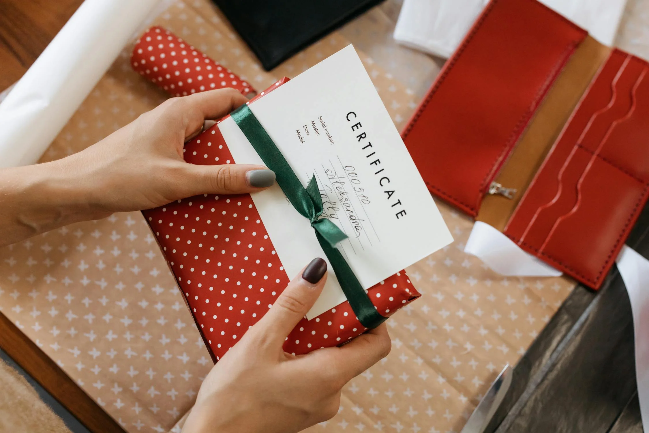 A hand holds a red polka-dotted gift with a green ribbon and certificate