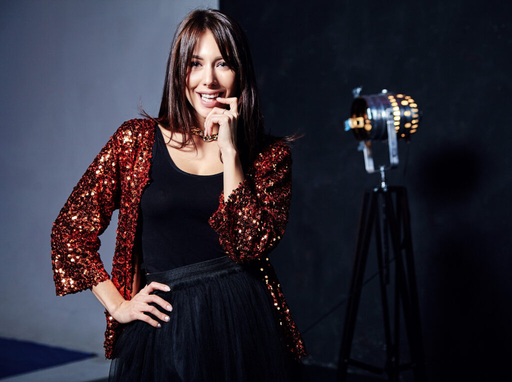 A stylish person poses in a sequined red jacket and black skirt, with a vintage spotlight in the background.