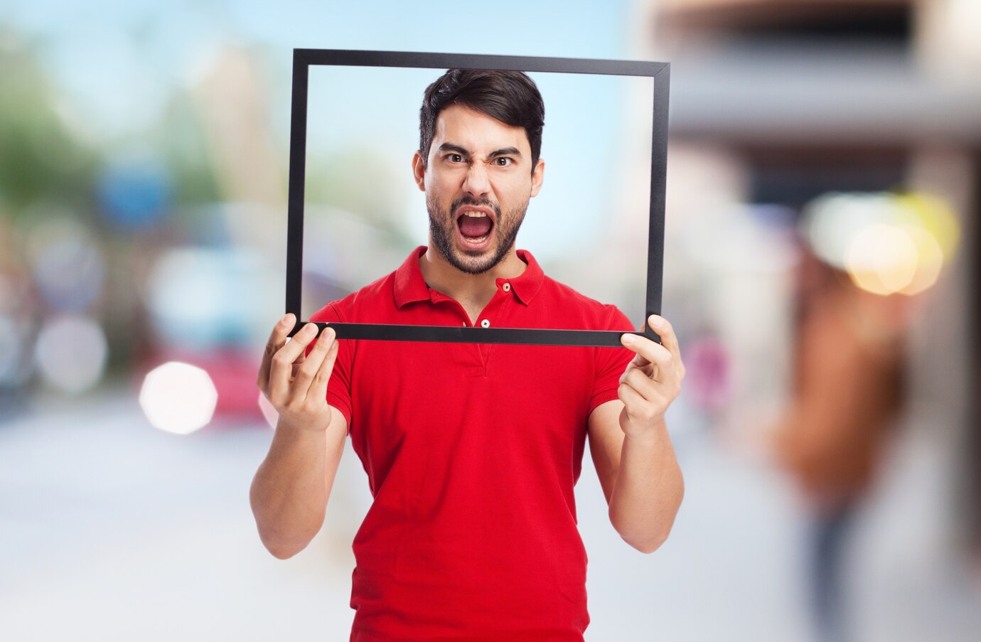A person wearing a red polo shirt holds a black frame in front of them, set against a blurred outdoor background.
