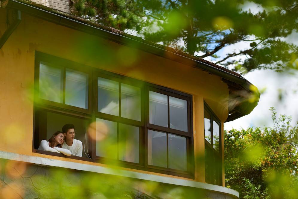 Lovers gaze out a large window of a cozy yellow home, surrounded by lush greenery