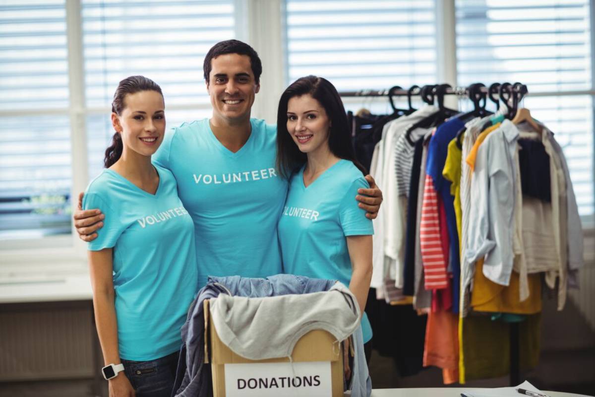 Three smiling volunteers in teal shirts stand by a donation cart in a clothing donation center.