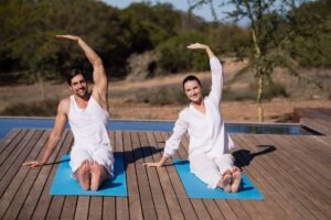 A man and a woman practicing yoga on mats outdoors by a pool, wearing white outfits, smiling.