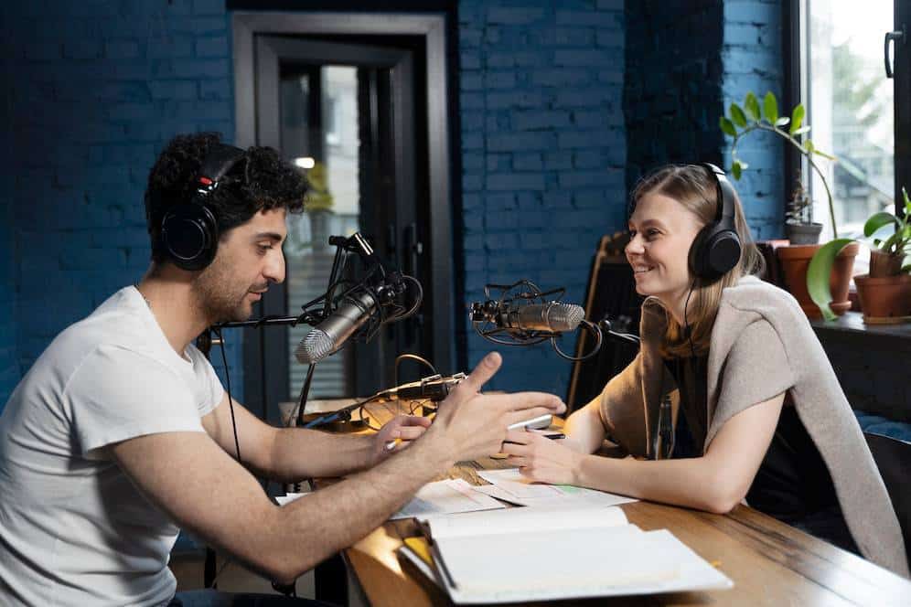 Two podcasters conversing enthusiastically over microphones in a studio with a blue brick wall background.