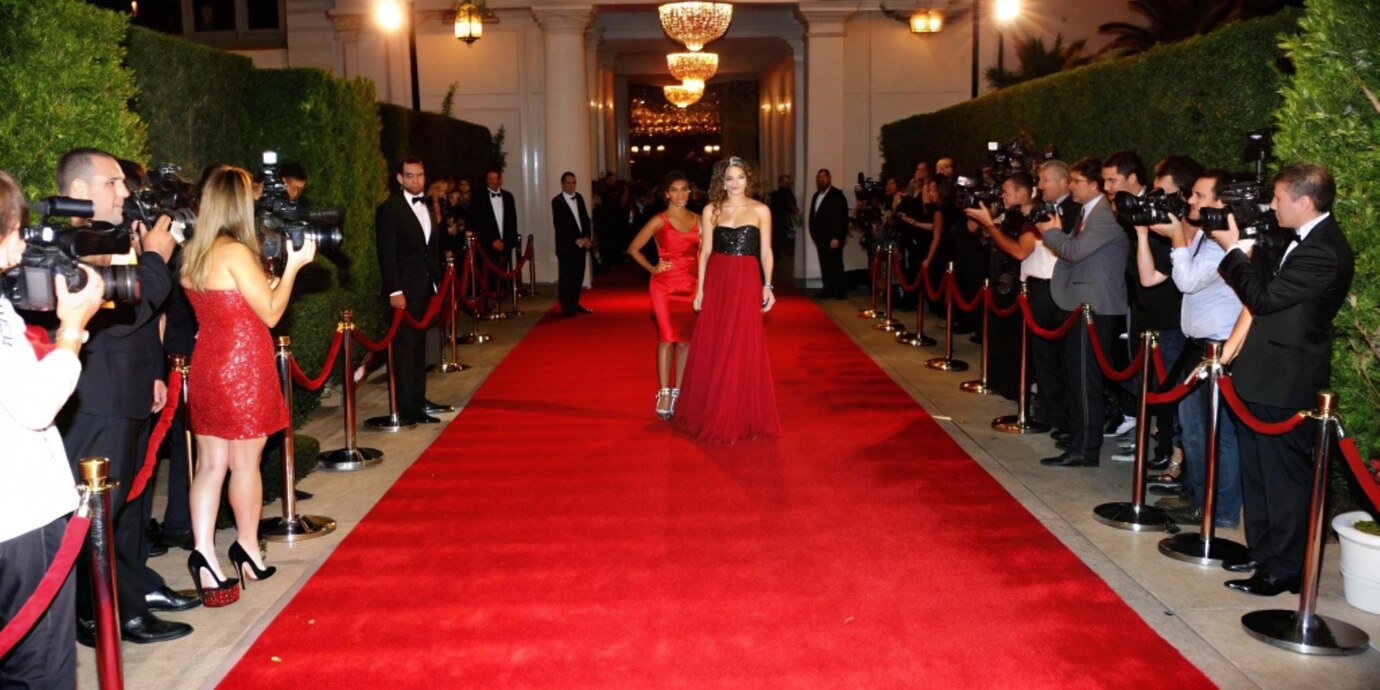 Two women in elegant dresses walk down a red carpet, surrounded by photographers capturing the event.