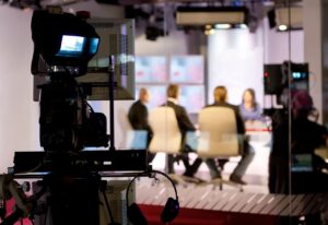 Professional video camera foreground focusing on a blurred panel of speakers in a TV studio.