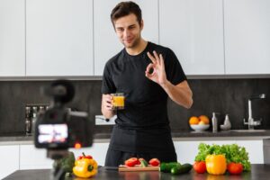 Young man in kitchen making an OK gesture while recording a cooking video, surrounded by fresh vegetables and holding orange juice.