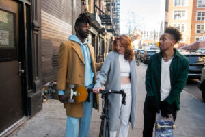Three young adults chatting on a city street, one holding a bicycle and another carrying a skateboard.