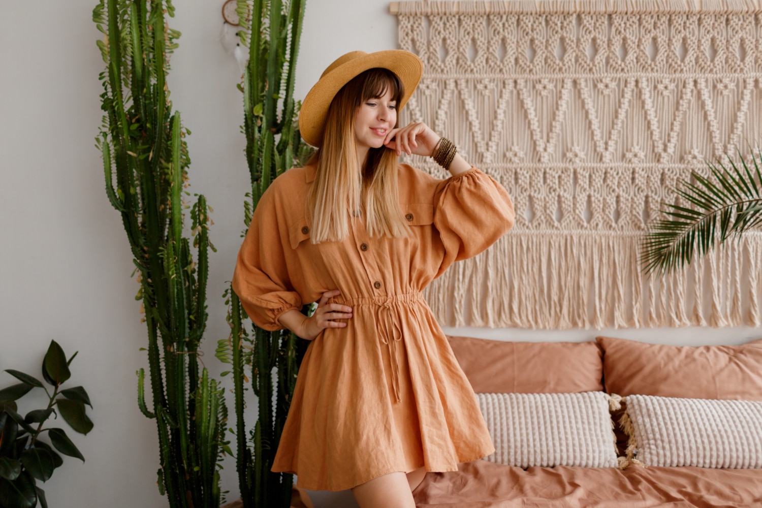 Woman in a straw hat and orange dress posing in a bohemian styled room with cacti and macrame decor.