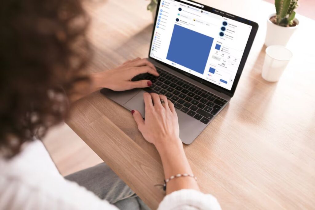 Woman using a laptop with social media interface displayed, at a wooden table with cacti and a ceramic cup nearby.