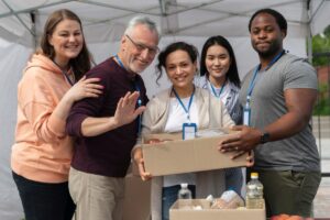 Group of five diverse volunteers wearing lanyards and smiling, holding a box of supplies at a community event.