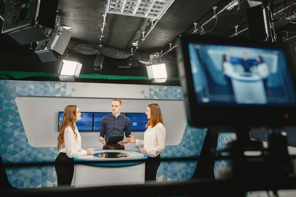 Three people discussing in a modern TV studio setup, with camera equipment visible.