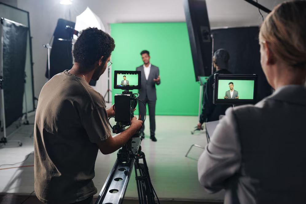 Two camera operators filming a man presenting in front of a green screen in a studio setting.