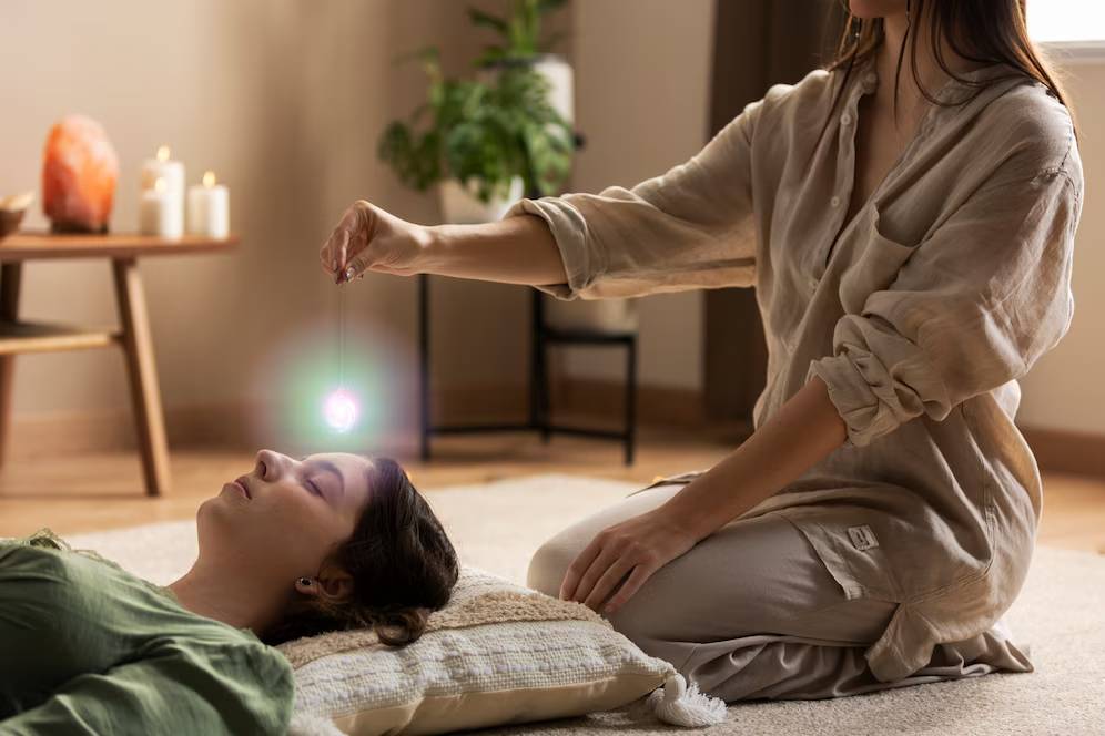 A woman performs a crystal healing session on a relaxed woman lying on a cushioned floor.