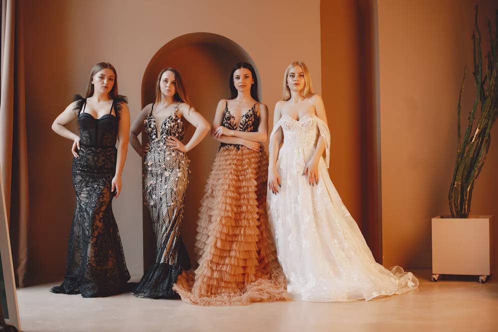 Four women in elegant evening gowns stand stylishly in a warm-hued setting