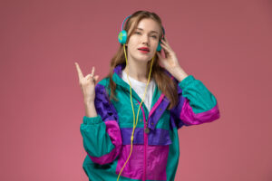 Young woman in colourful jacket listening to music with headphones, making peace sign, on pink background.