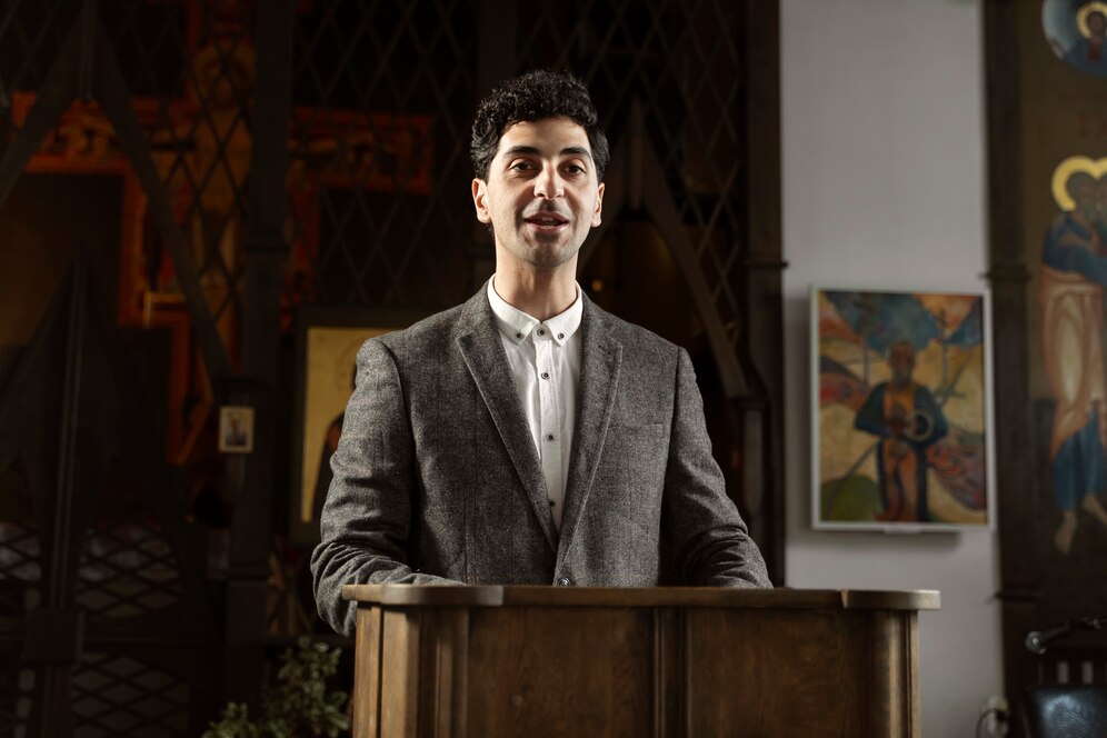 Man in grey blazer speaking at a wooden lectern, in a room adorned with religious art.
