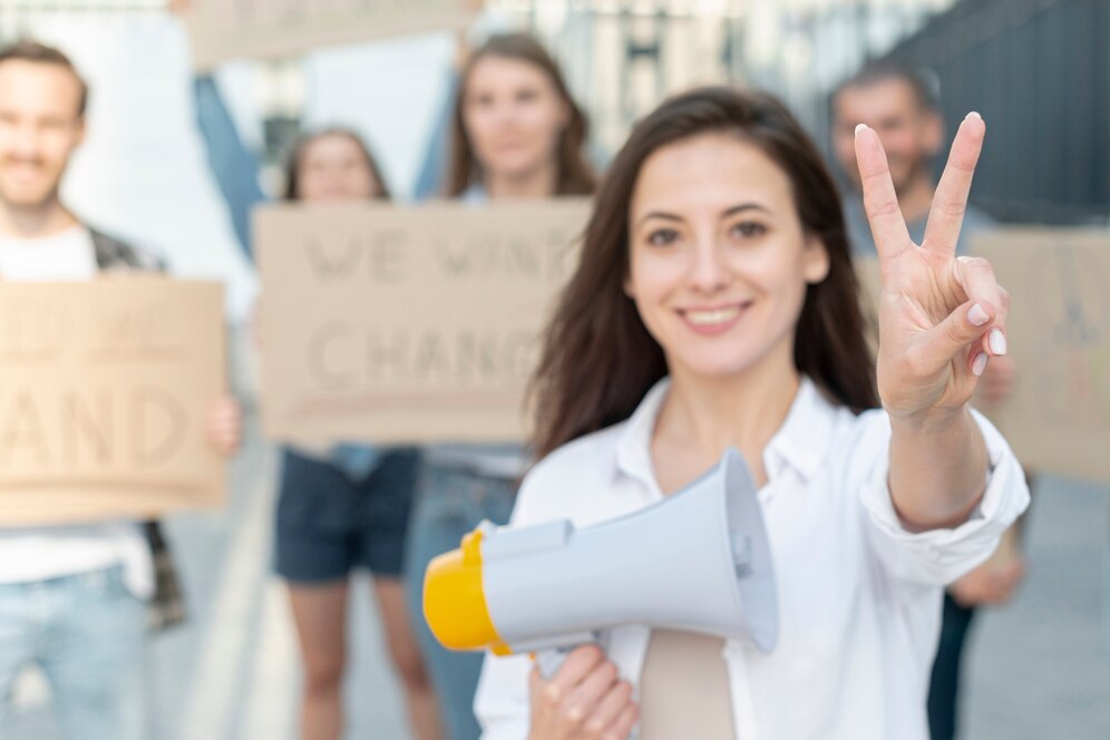 Young woman at a protest holding a megaphone and gesturing peace sign, with others holding signs in the background.