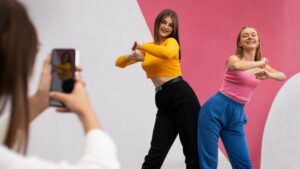 Two young women dancing and posing for a smartphone photo against a pink and white mural.