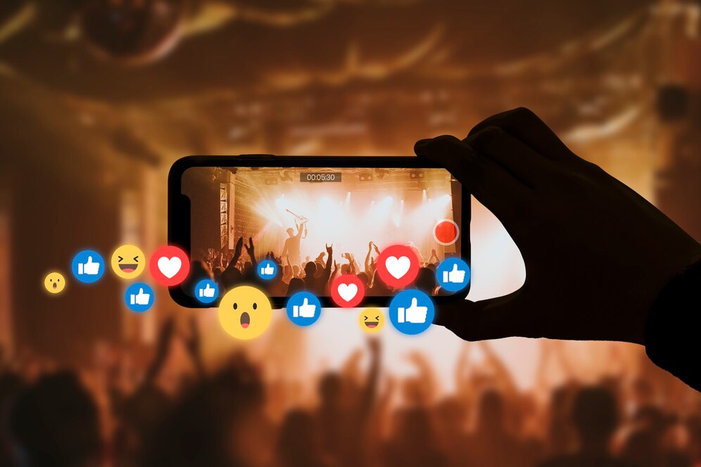 Hand holding a smartphone recording a live concert with emoji reactions displayed on the screen.