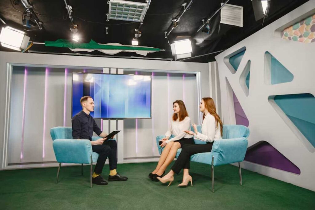 Three people in a modern television studio setting, two women speaking with a man holding a clipboard.