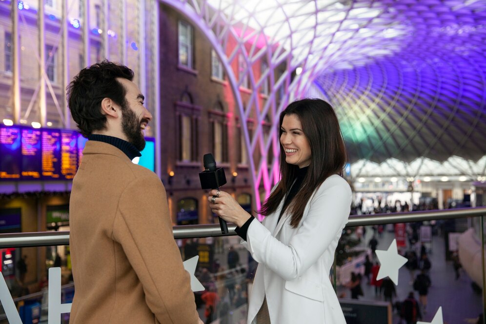 A man and a woman smiling and talking in a busy train station with an intricate roof structure.