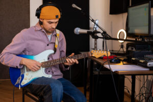 Man in orange beanie and headphones playing electric guitar in a home studio.