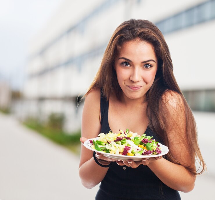 A young woman holding a plate of fresh salad, smiling at the camera, outdoor setting.