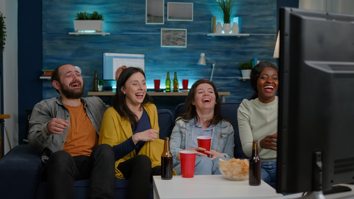 Four friends laughing and watching TV in a cozy living room, enjoying snacks and drinks.