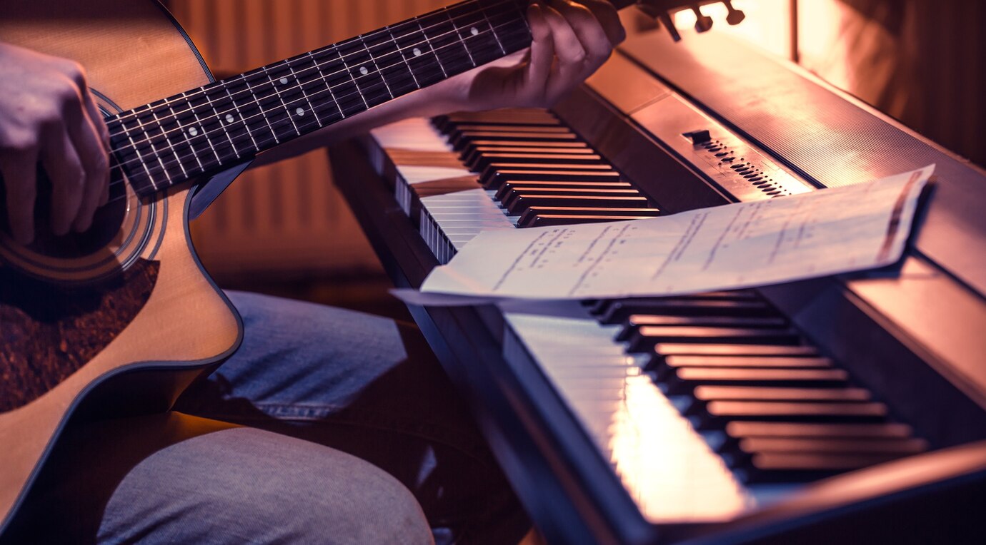 Person playing acoustic guitar beside a piano with sheet music on top in a dimly lit room.