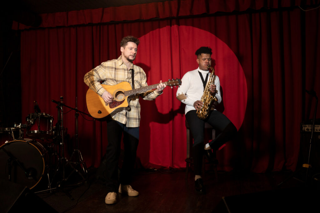 Two musicians performing on stage, one playing guitar and the other saxophone, with a red curtain backdrop.