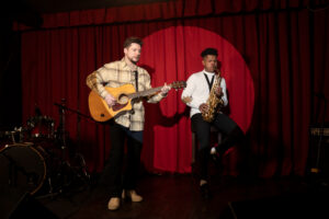 Two musicians performing on stage, one playing guitar and the other saxophone, with a red curtain backdrop.