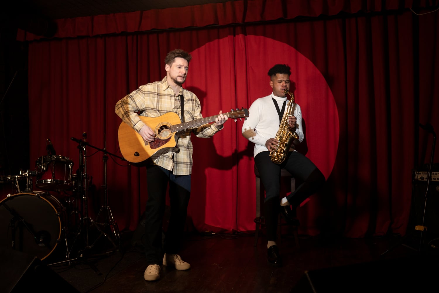 Two musicians performing on stage, one playing guitar and the other saxophone, with a red curtain backdrop.