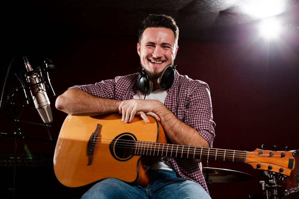 Joyful male musician laughing while holding a guitar in a recording studio, with microphone and studio light shining in the background.
