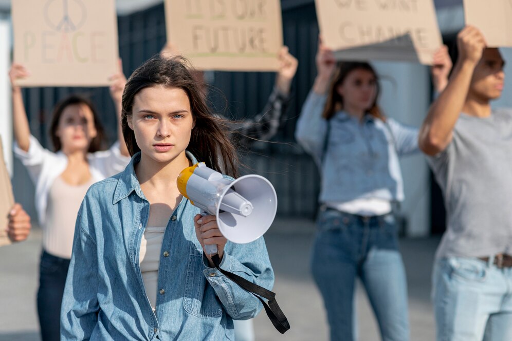 Young woman holding a megaphone at a peaceful protest with others holding signs in the background.