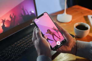 Person holding a smartphone displaying a dance video on a social media app, with a laptop and coffee cup in the background.
