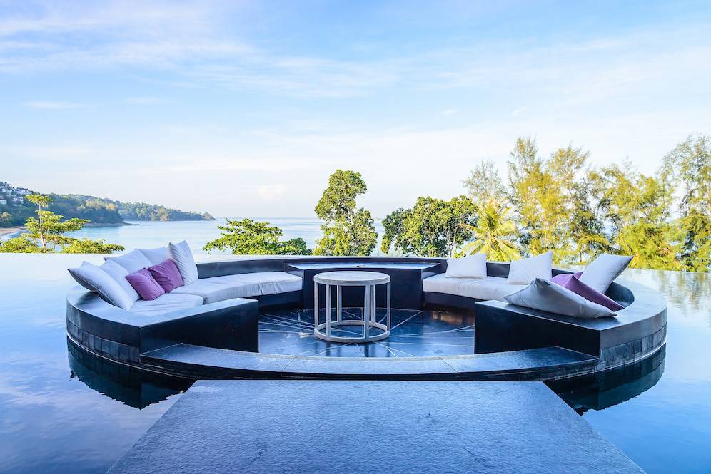 Outdoor circular lounge seating by an infinity pool overlooking a lush coastline under a clear blue sky.