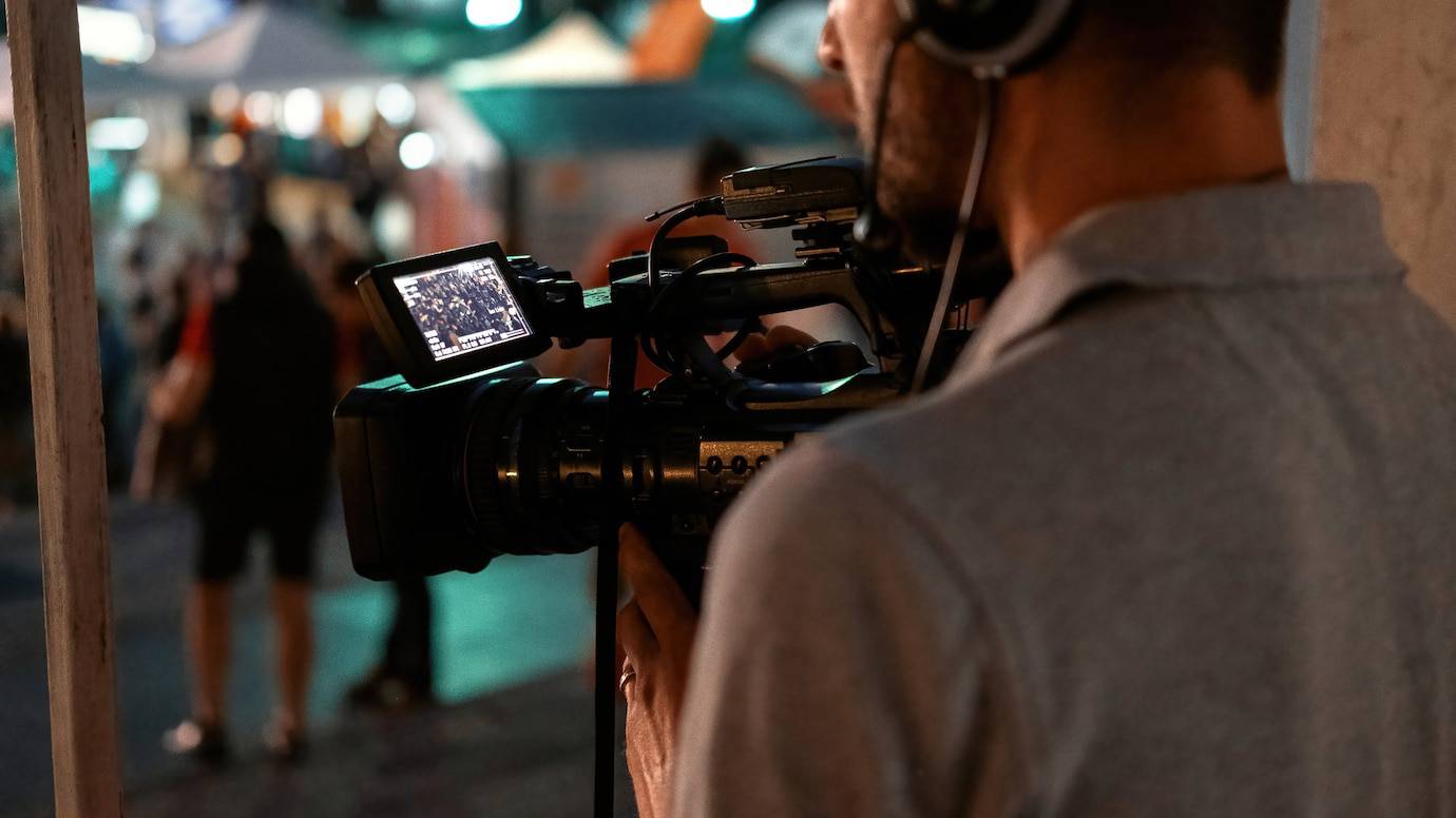 A cameraman recording a bustling street scene at night, viewed from behind his shoulder.