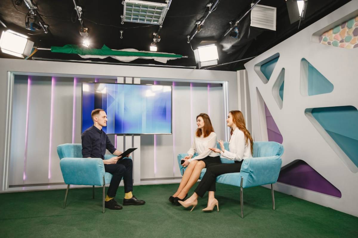 Two women in stylish outfits sit on a light blue couch, engaged in conversation with a person in the foreground