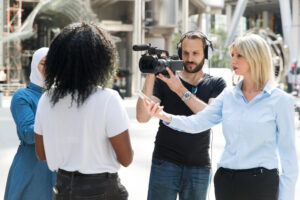 A film crew interviews a woman in a white t-shirt while a woman in a hijab
